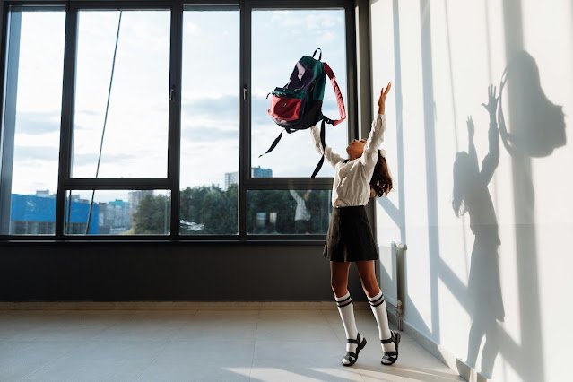 a little girl in school uniform throwing her backpack in the air in excitement. She is wearing a dark-colored skirt and a white blouse.