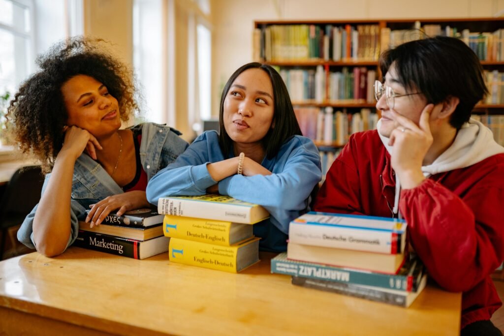 back to school college edition - 3 students chat together in the library surrounded by stacked books