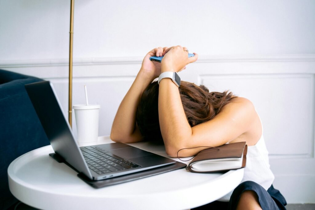 a woman sits procrastinating with her laptop on the table, head down on a small, white table, head resting between elbows, holding phone above her head with both hands