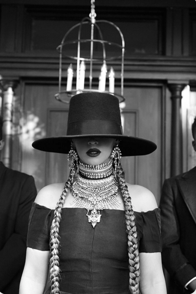 a still image from Beyonce's "Formation" video. Beyonce wears a wide brim hat that covers her eyes all the way to her nose. A little of her nose and all of her lips are showing from underneath the hat. She is wearing 2 French braids. The pic is black and white
