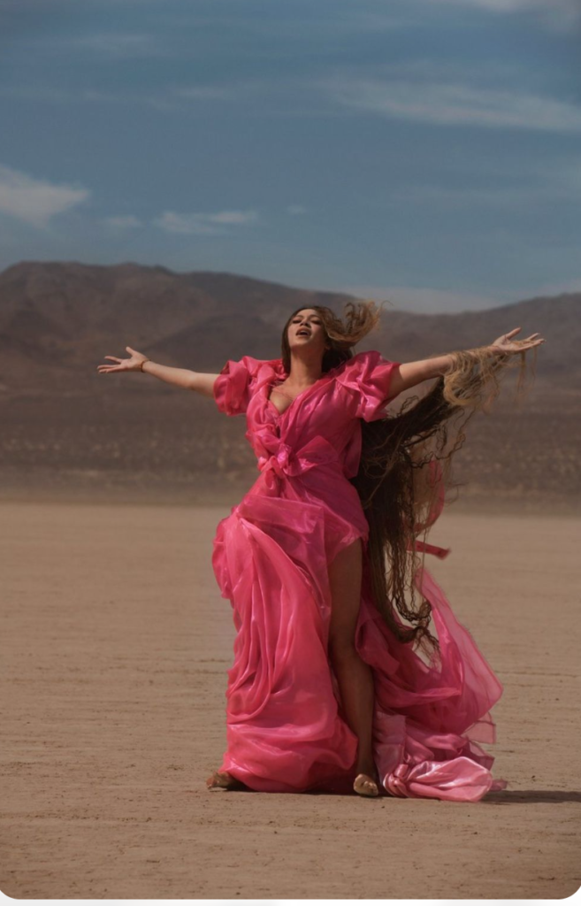 an image of Beyonce from her Lion King compilation. She is in the desert with a long pink gown and both arms out at shoulder level. Her face is toward the sky with her eyes closed.