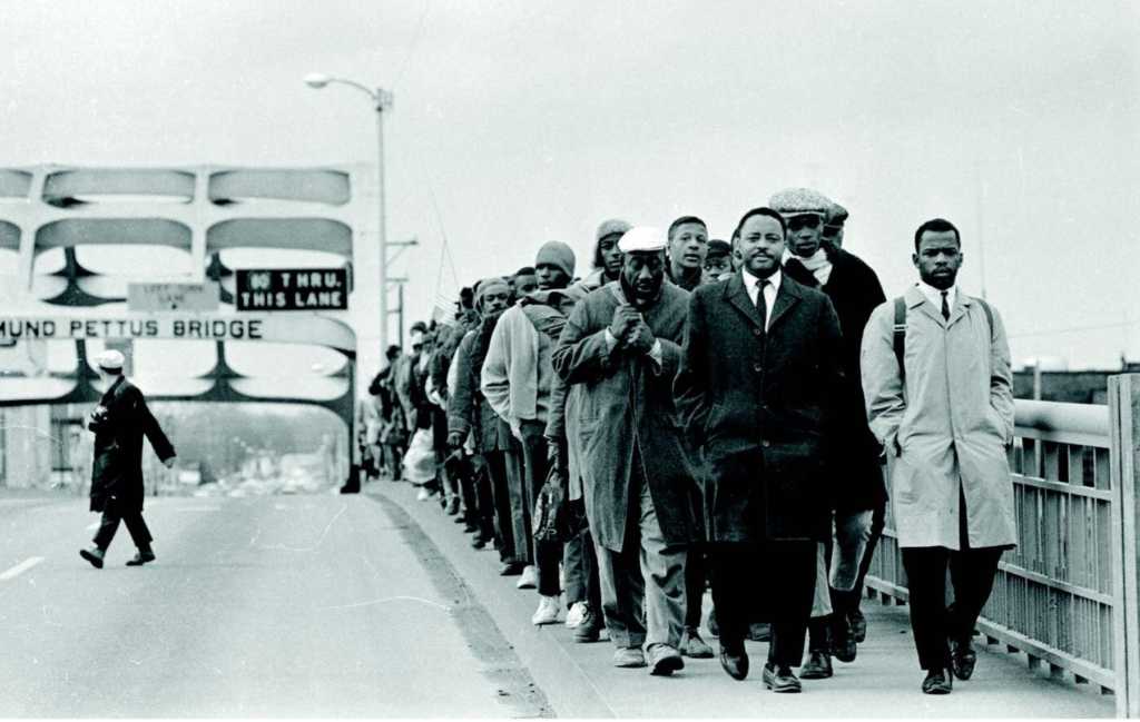 a black and white image of civil rights leaders crossing the Edmund Pettus Bridge led by John Lewis