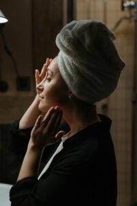 a woman stands in front of a mirror with her hair wrapped giving herself a facial