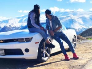 a couple outdoors - the woman sits on a white Camaro. the man leans against the car as they both take in the outdoors. Snow-capped mountains are in the background