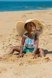 a precious toddler is making a sand castle on a beach. She wears an oversized sunhat and swimsuit. Aqua blue water is in the background