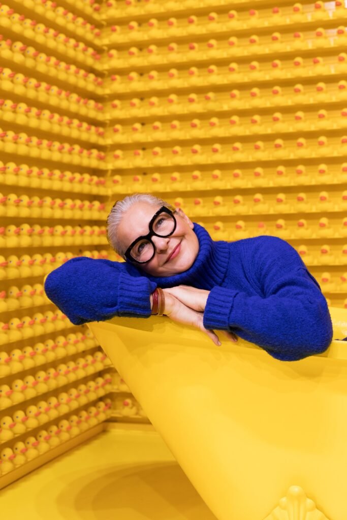 how to clear up skin over 40: Wall to wall rubber ducks and a bright yellow claw foot bathtub to match with this woman who is all smiles right in the middle. Her blue fashion stands out in the otherwise totally yellow room.