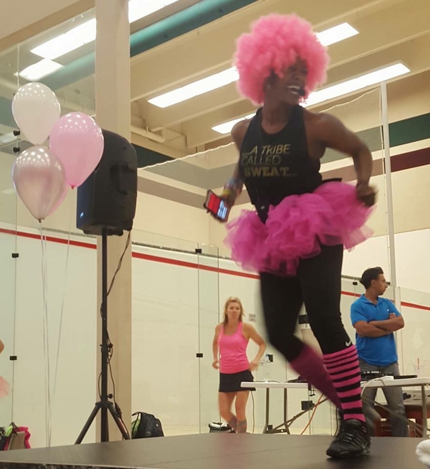 on stage teaching group fitness at lifetime fitness wearing a pink wig, blank tank top, black workout pants, pink tutu, and pink leg warmers surrounded by pink balloons for October's "Pink Out for Breast Cancer Awareness"