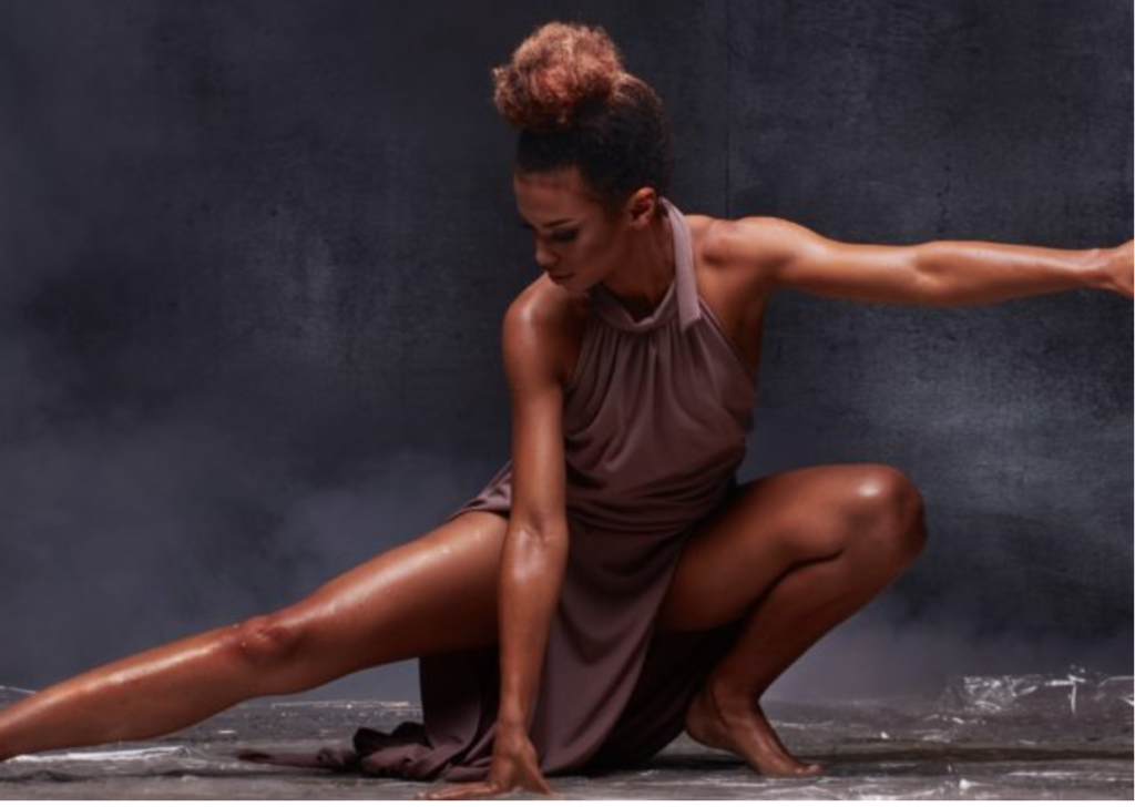 Black history through dance - an African American dancer (female) in a ballet pose squatted near floor, right leg extended, left arm extended at shoulder level. The lighting accenuates her toned arms and legs