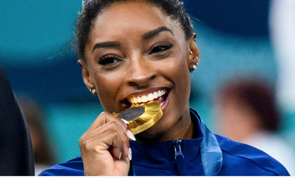 simone biles playfully bites her olympic gold medal as she smiles for the camera