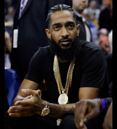 the late nipsey hussle sits courtside at an nba game wearing a black tee, gold jewelry and freshly done cornrows