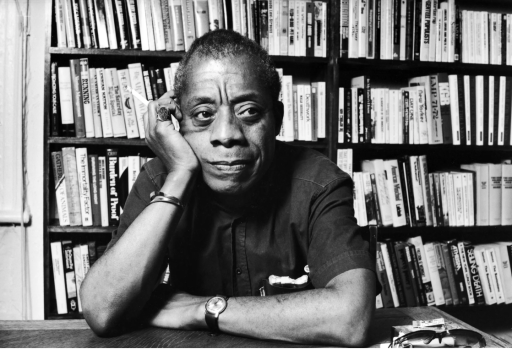Inspirational quotes for young black males -- Author and Civil Rights Activist, James Baldwin in black and white sitting in front of shelves of books