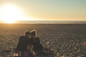 a couple sits on a blanket at the beach. Their heads touching. They are observing a beautiful sunset