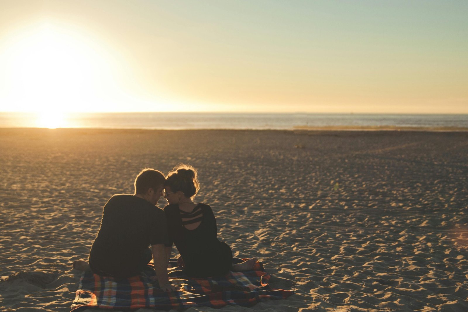a couple sits on a blanket at the beach. Their heads touching. They are observing a beautiful sunset