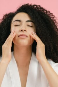 hydrating skincare ideas are plentiful. a young, fair-skinned woman with beautiful, hydrated skin poses with eyes closed and fingertips touching her face. the background is pink. she has long curly, voluptuous black hair