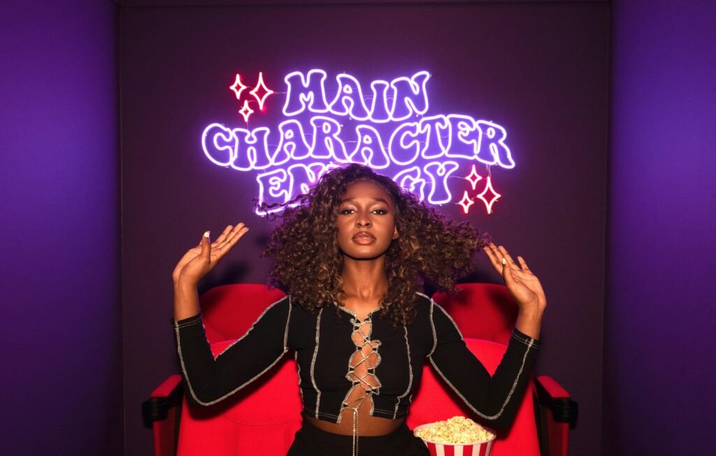 Baddie Aesthetic Quotes - a beautiful African American woman sits on a red couch with a bucket of buttered popcorn, on the wall behind her is a neon sign that says, "MAIN CHARACTER ENERGY."