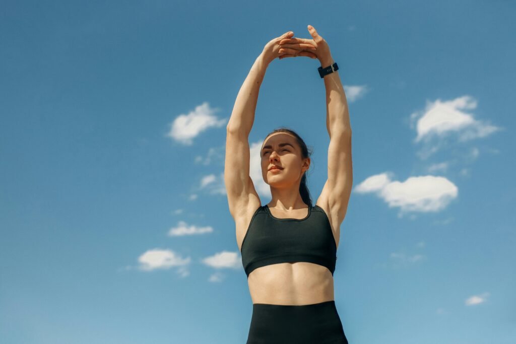 a woman with an athletic build stands outdoors with arms and hands outstretched above her head. She wears a sports bra and legs showing her toned midsection