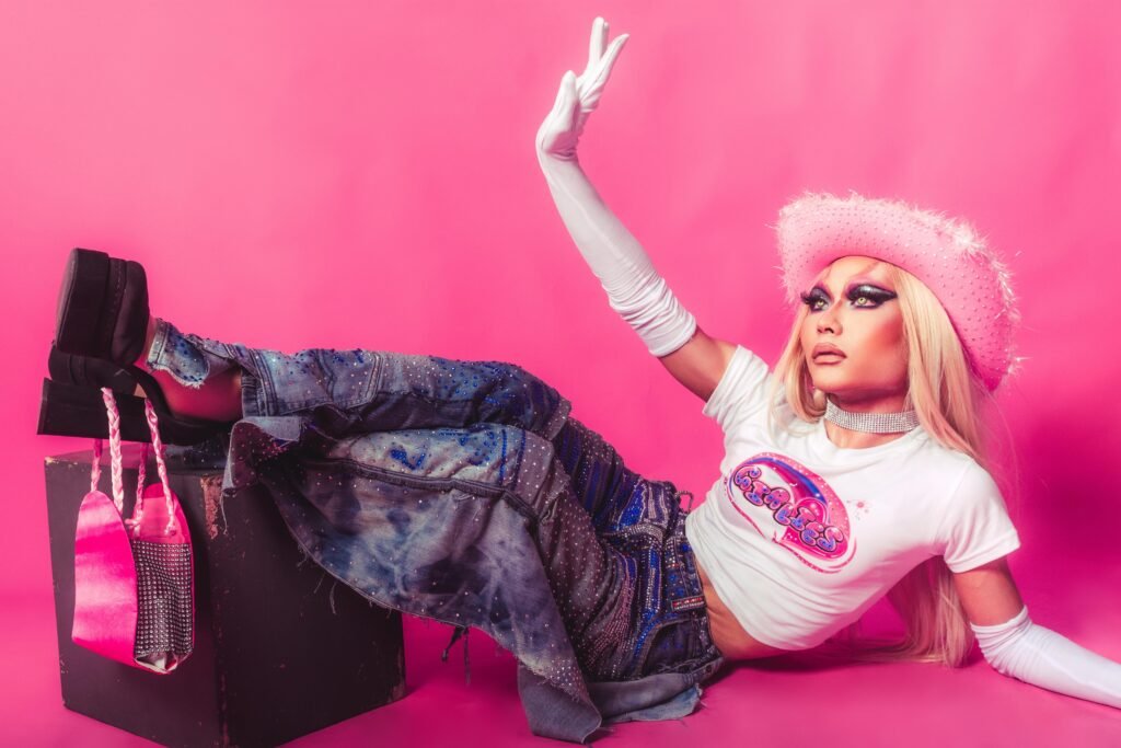 Baddie aesthetic quotes - a woman posing on the floor. the background is pink. she is wearing a pink hat, pink tee, jeans, platform heels, a pink purse hangs from 1 heel, and full-length white gloves. she has full makeup on with colored contacts and a long, straight hair and blond wig