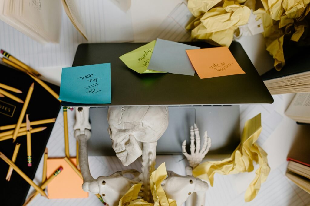 get your life together - an image of someone who has procrastinated either himself or herself into a skeleton. The skeleton's head and arms are on a desk and laptop surrounded by crumpled up papers, post it notes, and broken pencils. A mess!