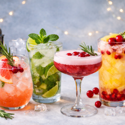 A realistic photo of four refreshing non alcoholic mocktails on ice, garnished with citrus slices, mint, rosemary, and cranberries, arranged on a light surface with fresh fruit and cocktail tools in the background.